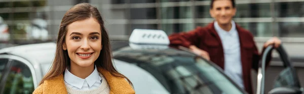 Foco seletivo da mulher sorrindo para a câmera com motorista de táxi no fundo, tiro panorâmico — Fotografia de Stock