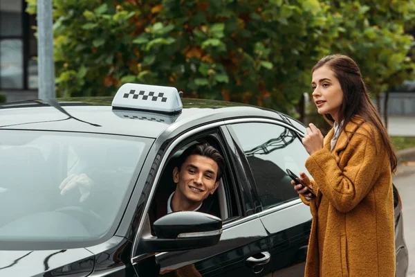 Молодая женщина держит смартфон и разговаривает с улыбающимся таксистом — стоковое фото