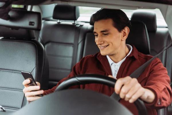 Uomo sorridente guardando smartphone durante la guida di auto — Foto stock