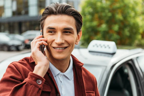 Focus sélectif d'un homme souriant parlant sur smartphone avec taxi en arrière-plan — Photo de stock