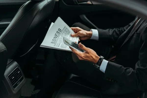 Vista recortada del empresario sosteniendo periódico y teléfono inteligente en taxi - foto de stock
