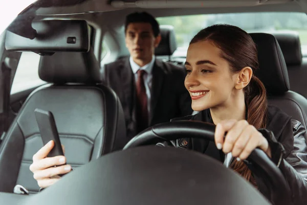 Taxista sonriente usando teléfono inteligente con hombre de negocios en segundo plano - foto de stock