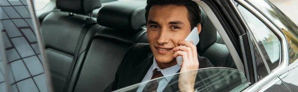 Sonriente hombre de negocios hablando en el teléfono inteligente en el asiento trasero del taxi, plano panorámico - foto de stock