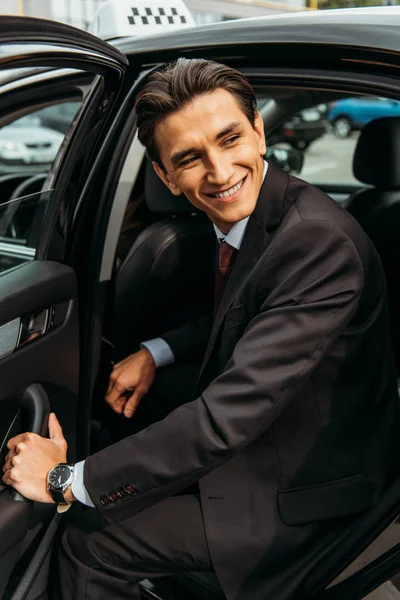 Un hombre de negocios sonriente bajándose del taxi y mirando hacia otro lado - foto de stock