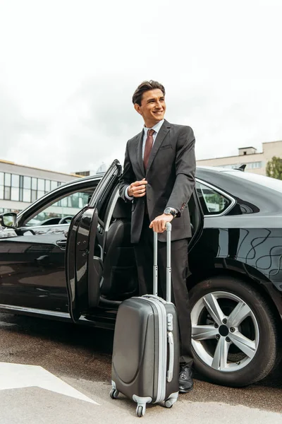 Smiling businessman with suitcase standing by taxi — Stock Photo