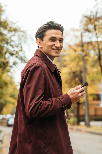 Sonriente hombre sosteniendo el teléfono inteligente y mirando a la cámara en la calle - foto de stock