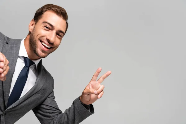 Bonito e sorridente empresário em terno mostrando sinal de paz isolado no cinza — Fotografia de Stock