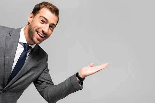 Bonito e sorridente empresário em terno apontando com a mão isolada em cinza — Fotografia de Stock