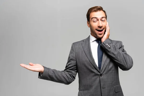 Hombre de negocios guapo y sorprendido en traje apuntando con la mano aislada en gris - foto de stock