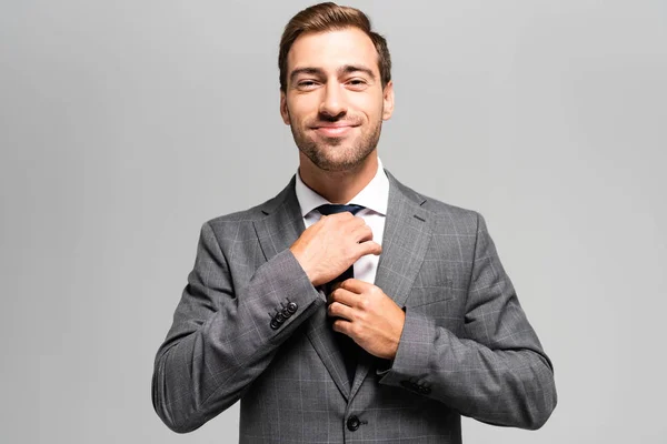 Handsome and smiling businessman in suit touching tie isolated on grey — Stock Photo