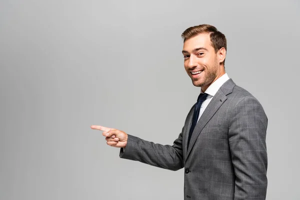 Hombre de negocios sonriente y guapo en traje apuntando con el dedo aislado en gris - foto de stock