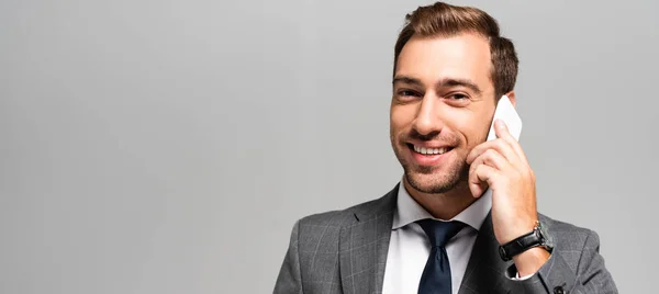 Panoramic shot of handsome and smiling businessman in suit talking on smartphone isolated on grey — Stock Photo