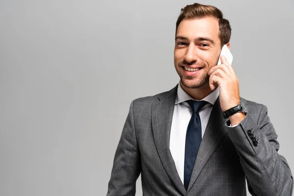 Bonito e sorridente empresário em terno falando no smartphone isolado em cinza — Fotografia de Stock