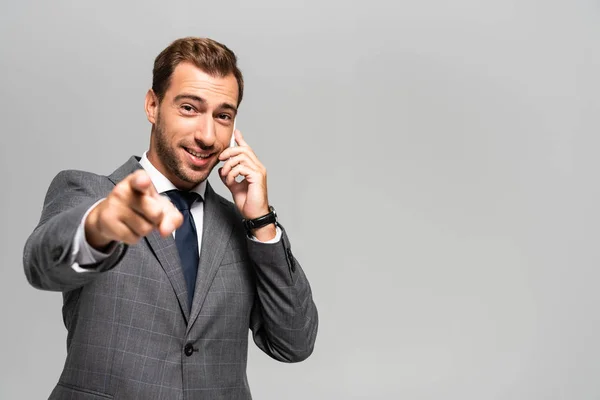 Hombre de negocios guapo y sonriente en traje hablando en el teléfono inteligente y señalando con el dedo aislado en gris - foto de stock