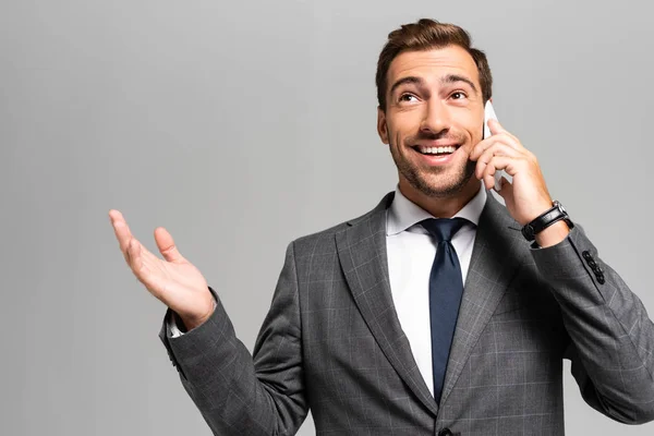 Bonito e sorridente empresário de terno com mão estendida falando em smartphone isolado em cinza — Fotografia de Stock