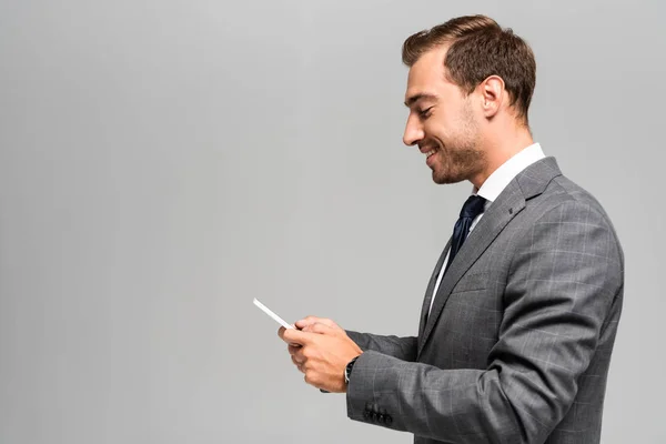 Seitenansicht des lächelnden und gut aussehenden Geschäftsmannes im Anzug mit Smartphone isoliert auf grau — Stockfoto