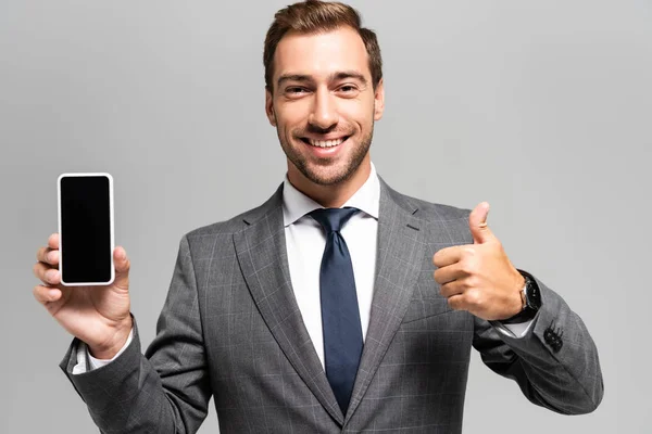 Hombre de negocios sonriente en traje sosteniendo el teléfono inteligente y mostrando como signo aislado en gris - foto de stock