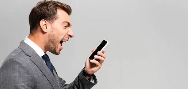 Plano panorámico de hombre de negocios guapo y enojado en traje gritando a teléfono inteligente aislado en gris - foto de stock