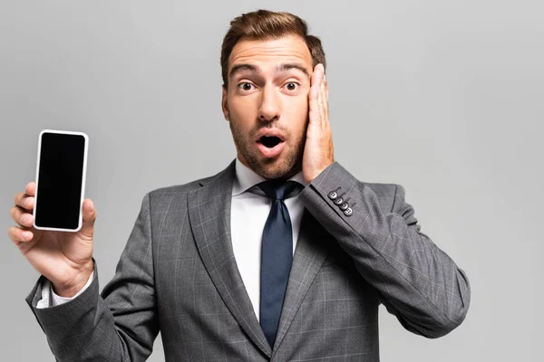 Handsome and shocked businessman in suit holding smartphone isolated on grey — Stock Photo