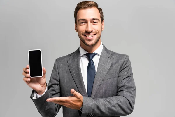 Hombre de negocios guapo y sonriente en traje apuntando con la mano al teléfono inteligente aislado en gris - foto de stock