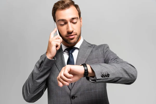 Красивый бизнесмен в костюме разговаривает по смартфону и смотрит на часы, изолированные на сером — стоковое фото