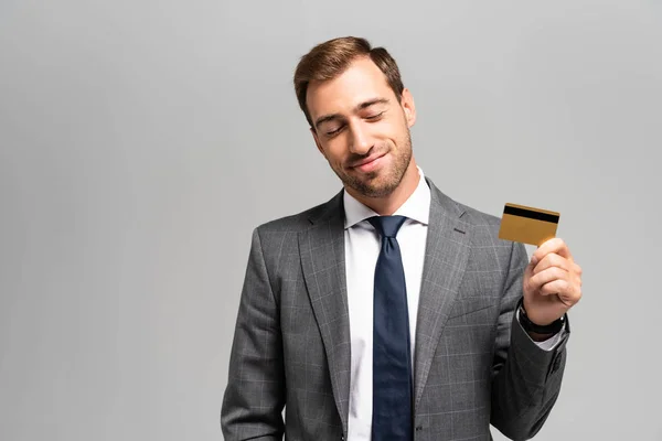Красивый и улыбающийся бизнесмен в костюме, держащий кредитную карту изолированной на серый — стоковое фото