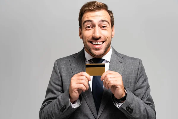 Hombre de negocios guapo y sonriente en traje con tarjeta de crédito aislado en gris - foto de stock