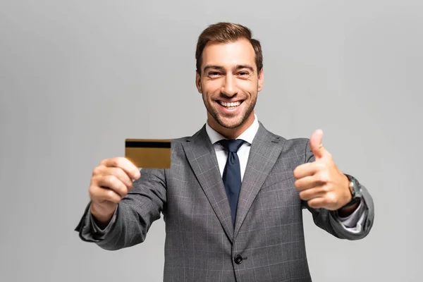 Beau et souriant homme d'affaires en costume tenant la carte de crédit et montrant comme signe isolé sur gris — Photo de stock
