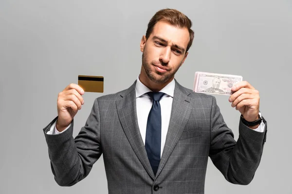 Гарний і сумний бізнесмен у костюмі, що тримає кредитну картку і доларові банкноти ізольовані на сірому — стокове фото