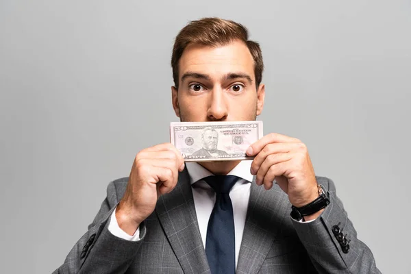 Apuesto hombre de negocios en traje que oscurece la cara con billete de dólar aislado en gris - foto de stock