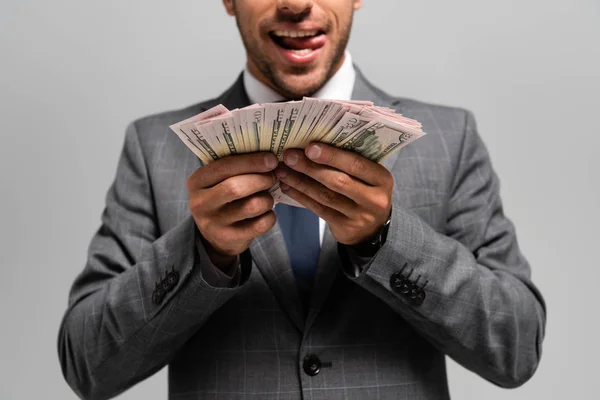 Cropped view of businessman in suit holding dollar banknotes and licking lips isolated on grey — Stock Photo