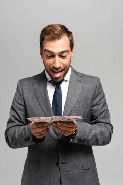 Красивый и улыбающийся бизнесмен в костюме держит долларовые купюры изолированные на сером — стоковое фото