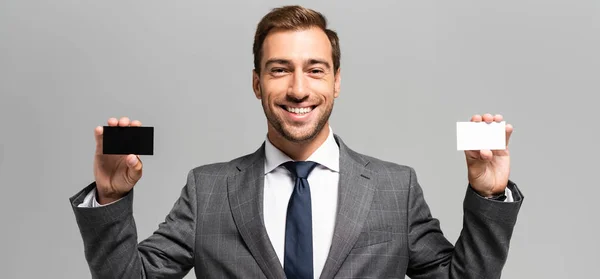 Панорамный снимок красивого и улыбающегося бизнесмена в костюме держащего визитки изолированные на сером — стоковое фото