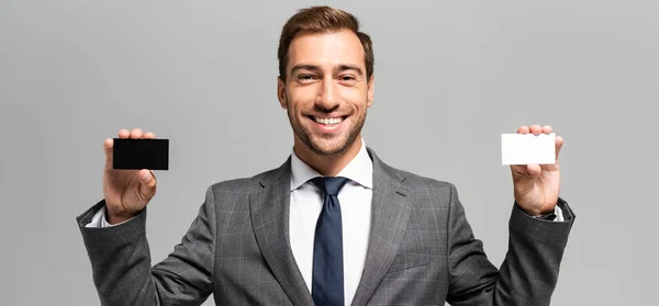 Panoramic shot of handsome and smiling businessman in suit holding business cards isolated on grey — Stock Photo