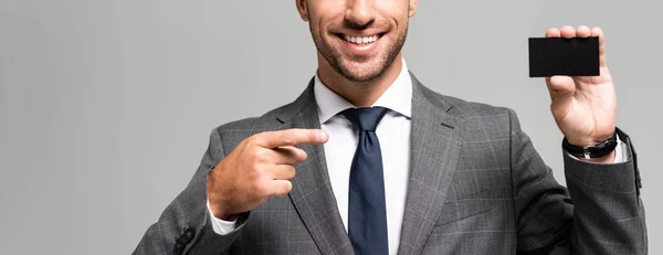 Panoramaaufnahme eines lächelnden Geschäftsmannes im Anzug, der mit dem Finger auf eine auf Grau isolierte Visitenkarte zeigt — Stockfoto