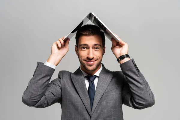 Hombre de negocios guapo y sonriente en traje que sostiene el ordenador portátil por encima de la cabeza aislado en gris - foto de stock