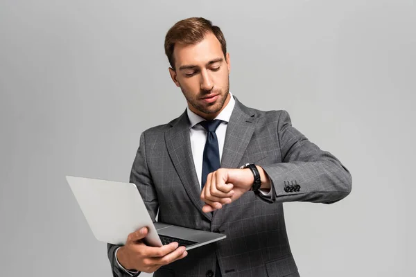 Handsome businessman in suit holding laptop and looking at wristwatch isolated on grey — Stock Photo