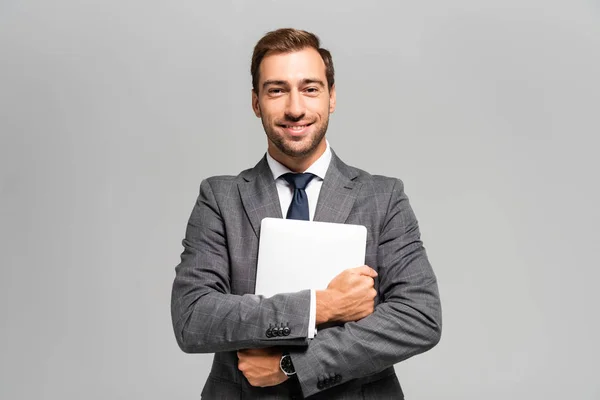 Красивый и улыбающийся бизнесмен в костюме держит ноутбук изолированным на сером — стоковое фото