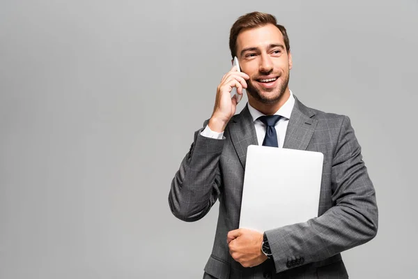 Hombre de negocios guapo y sonriente en traje que sostiene el ordenador portátil y habla en el teléfono inteligente aislado en gris - foto de stock