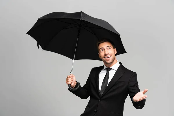 Hombre de negocios guapo y sonriente con la mano extendida en traje con paraguas aislado en gris - foto de stock