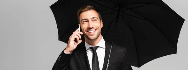 Panoramaaufnahme eines gut aussehenden und lächelnden Geschäftsmannes, der mit seinem Smartphone spricht und einen Regenschirm isoliert auf grau hält — Stockfoto