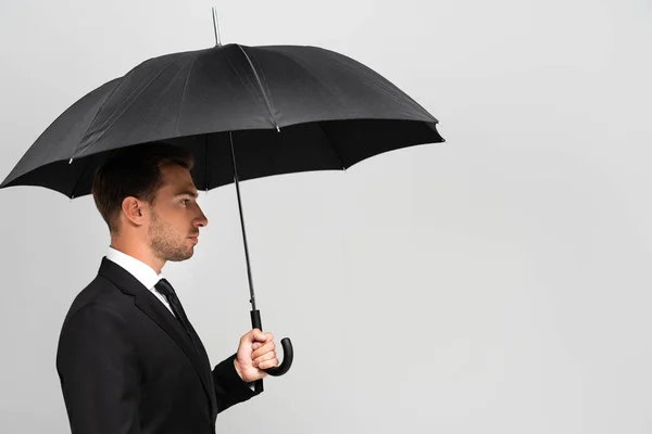 Vista lateral del hombre de negocios guapo en traje con paraguas aislado en gris - foto de stock