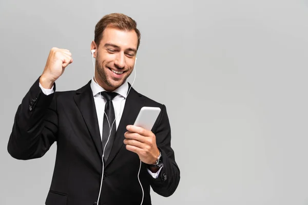 Bonito e sorridente empresário de terno com fones de ouvido usando smartphone e mostrando sim gesto isolado em cinza — Fotografia de Stock