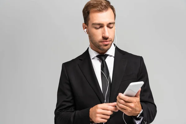 Handsome businessman in suit with earphones using smartphone isolated on grey — Stock Photo