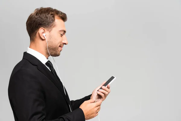 Vista lateral del hombre de negocios guapo y sonriente en traje con auriculares usando teléfono inteligente aislado en gris - foto de stock