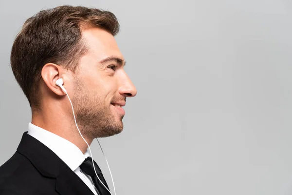 Side view of handsome and smiling businessman in suit listening music with earphones isolated on grey — Stock Photo