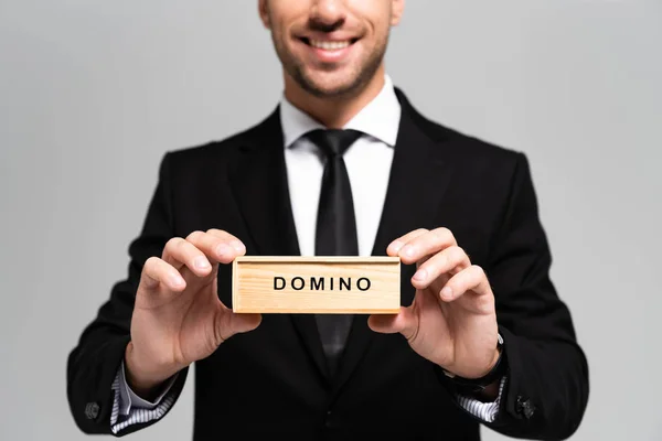 Vista ritagliata di uomo d'affari sorridente in giacca e cravatta con scatola di legno con scritte domino isolato su grigio — Foto stock