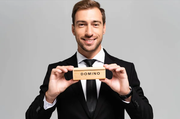 Homme d'affaires souriant en costume tenant boîte en bois avec lettrage domino isolé sur gris — Photo de stock