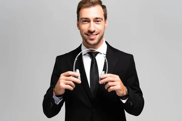 Sonriente hombre de negocios con traje sosteniendo auriculares aislados en gris - foto de stock