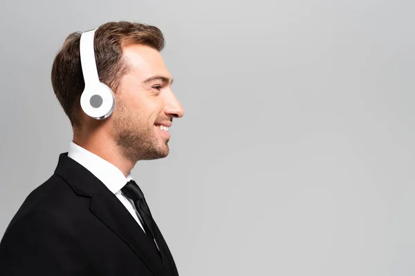 Vista lateral del hombre de negocios sonriente en traje con auriculares escuchando música aislada en gris - foto de stock